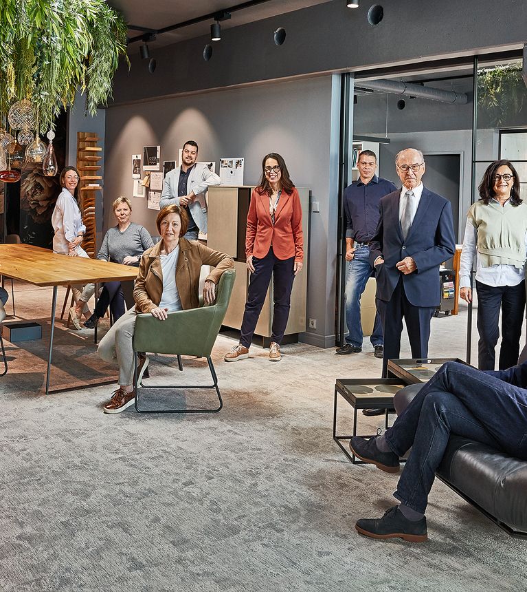 Eine Gruppe von zwölf Personen in einem modernen Büro, die um einen Tisch herum sitzen und stehen, mit Pflanzen und stilvollen Möbeln im Hintergrund.